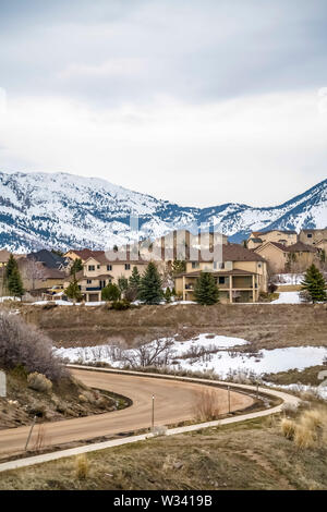 Paved road curving through snowy hill with multi storey homes in the distance. A view of snow covered mountain against cloudy sky can also be seen on Stock Photo