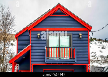 Close up of home exterior with vibrant blue wall red frames and small balcony. Cloudy sky and mountain covered with snow in winter cna be seen in the Stock Photo