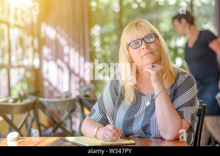 Portrait of beautiful plus size middle age woman working and making notes to her diary on sunny terrace of cafe. Elegant middle aged lady posing with Stock Photo