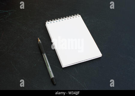 Open a blank notepad on the spiral on a black board background with black pen. Stock Photo