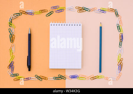 Notebook in cage with a pen and pencil in a frame of colored paper clips on a double background - beige and orange. Concept back to school. Stock Photo