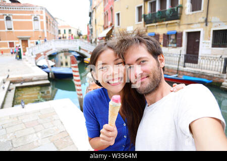 Couple in Venice, eating Ice cream taking selfie self-portrait photo on vacation travel in Italy. Smiling happy Asian woman and Caucasian man in love having fun eating italian gelato food outdoors. Stock Photo