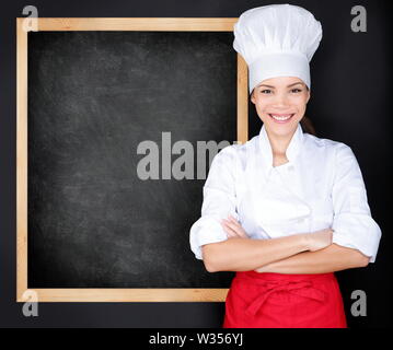 Chef showing menu blackboard. Woman in front of blank menu blackboard. Happy female chef, cook or baker by empty chalkboard menu display wearing chef whites uniform and hat Stock Photo