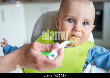 Portrait Of Smiling Baby Boy At Home In High Chair Being Fed Solid Food By Mother With Spoon Stock Photo
