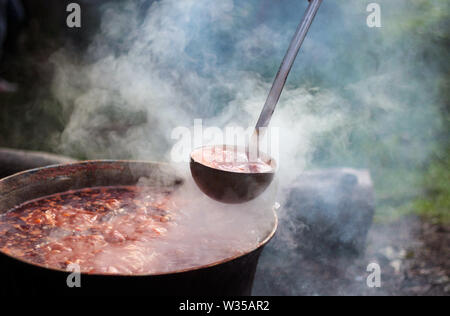 Outdoor rural summer scene where above the orange flames the traditional Hungarian goulash is boiling in a cauldron with a ladle Stock Photo