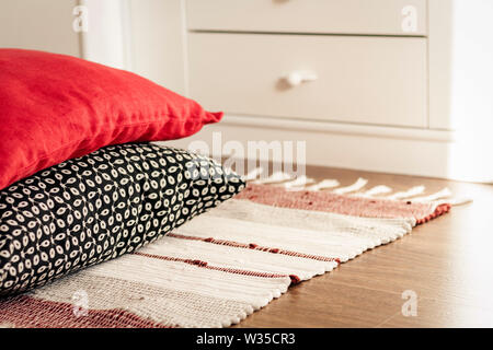 Cozy home interior decor, decorative pillows in front of white nightstand near a multi-colored rug background Stock Photo