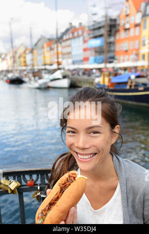 Woman eating traditional danish fast food snack hot dog. Girl enjoying hot dogs outside in Nyhavn waterfront canal street of Copenhagen, Denmark. Stock Photo