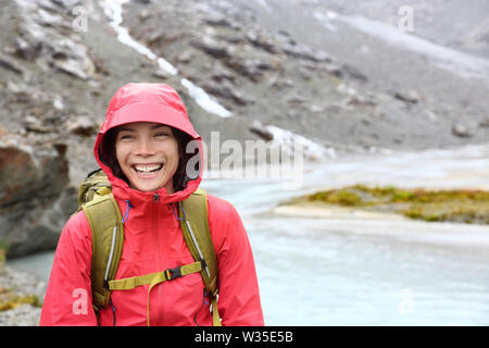 Hiker woman hiking with backpack in rain on trek living healthy active lifestyle. Smiling cheerful girl walking on hike in beautiful mountain nature landscape while raining in Swiss alps, Switzerland. Stock Photo