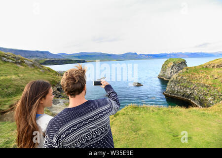 Tourists on travel taking photo with smartphone on Iceland. Happy couple sightseeing taking pictures using smart phone visiting Arnarstapi, Snaefellsnes, Iceland. Stock Photo