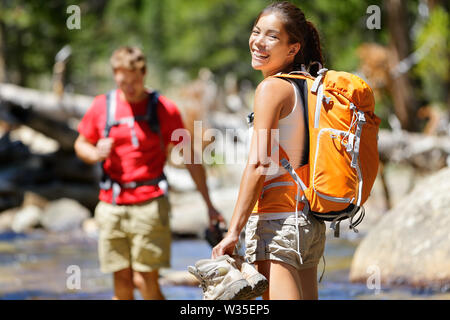 Hiking friends having fun crossing river in forest. Young happy adults barefoot walking in water with wet feet on an adventure trip hike in nature. Stock Photo