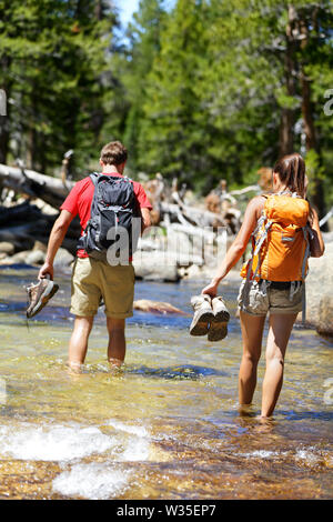 Hikers group walking barefoot crossing river in forest. Adventure people on hike hiking in nature holding shoes and boots to cross with wet feet. Stock Photo