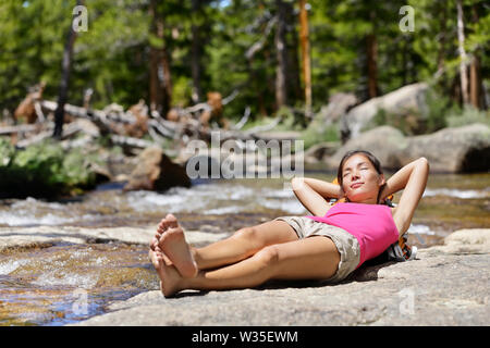 Hiking woman relaxing alone sleeping by river creek in nature. Tired hiker resting lying down outdoors taking a break from hike. Young Asian woman in forest in Yosemite national park, California, USA. Stock Photo
