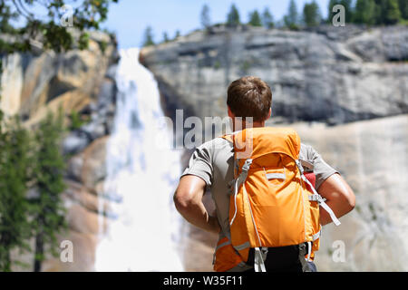 Hiker hiking with backpack looking at waterfall in Yosemite park in beautiful summer nature landscape. Portrait of male adult back standing outdoor. Stock Photo