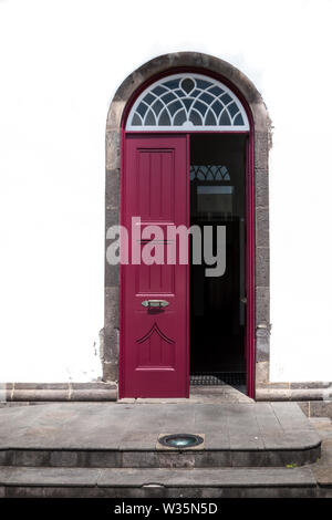 Half opened burgundy wooden traditional door of a house with bright white facade. White grating and an arc on the top. Povoacao, Sao Miguel, Azores Is Stock Photo