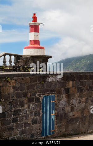 Stone wall and fence on the promenade along the Atlantic ocean. On the corner small red-white lighthouse. Blue sky with white clouds. Povoacao, Sao Mi Stock Photo