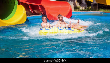 Family just after riding down the water park slide in yellow inflatable ring surrounded by water splashes. Father and son are enjoying summer weekend Stock Photo