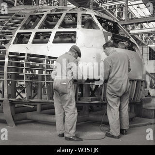 1950s, historical, aviation, two male mechanics or engineers in overalls working on the exterior of an aircraft cockpit inside an aerospace factory or hanger, England, UK. Stock Photo