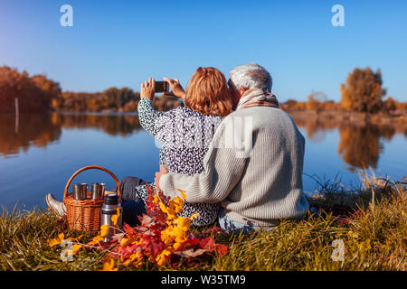 Senior couple taking selfie on phone while having picnic by autumn lake. Happy man and woman enjoying nature and hugging Stock Photo