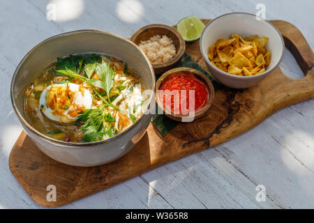 Soto ayam, a yellow spicy chicken soup with boiled egg, parsley and bawang goreng (fried shallots). Served with lime, spicy sambal sauce, potato chips Stock Photo