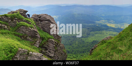 One man in a hat climbing on rocks on Pikuy mount against the valley among majestic green mountain hills covered in lush grass and forest. Cloudy summ Stock Photo