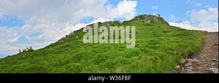 Tiny figures of several man standing on the peak of Pikuy mount against the blue sky with white clouds and lush grass. Cloudy summer day. Ukraine, Lvi Stock Photo