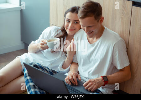 Dark-eyed wife drinking coffee while husband working on laptop Stock Photo
