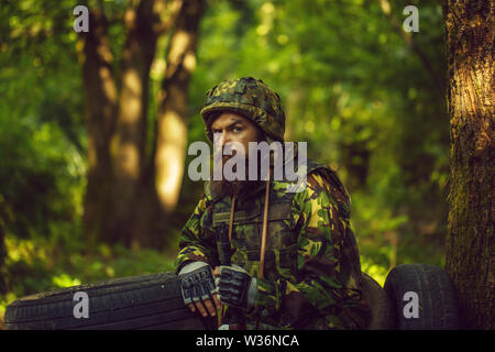Young soldier wirh bearded dusty tired face in military camouflage uniform with hanging camera and rifle in hand sitting on guard near tree and black Stock Photo