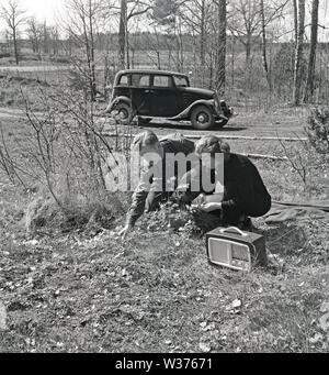 Lifestyle in the 1950s. A young couple having a car drive in the countryside,  has stopped  to pick some flowers on the roadside. They have brought a portable radio recieiver along and listens to it. Sweden 1952.  Kristoffersson Ref BF77-5. Stock Photo