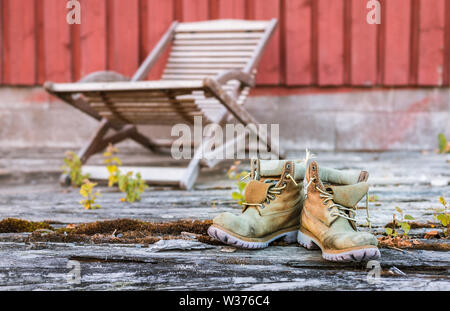 Pair of worn work shoes in front of wooden lounge chair and red wooden wall Stock Photo