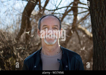 Outdoors portrait of mature senior man with grey hair and beard in dark leather jacket standing near the tree on blue sky background in early spring. Stock Photo
