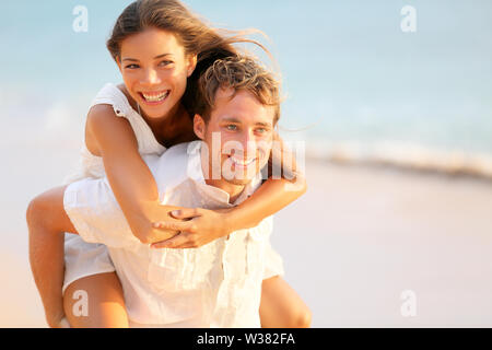 Lovers couple in love having fun dating on beach portrait. Beautiful healthy young adults girlfriend piggybacking on boyfriend hugging happy. Multiracial dating or healthy relationship concept. Stock Photo
