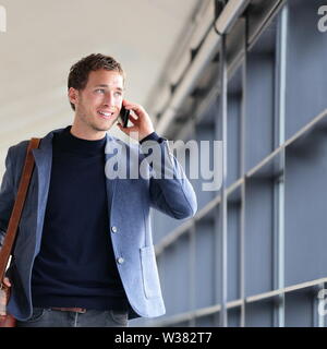 Urban business man talking on smart phone traveling walking in full body length inside in airport. Casual young businessman wearing suit jacket and shoulder bag. Handsome male model in his 20s. Stock Photo