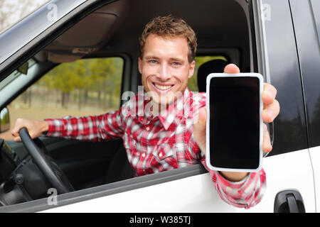 Smartphone man driving car showing app on screen display smiling happy. Male driver using 4g apps showing blank empty touchscreen sitting in drivers seat. Focus on model and on screen for copy space. Stock Photo