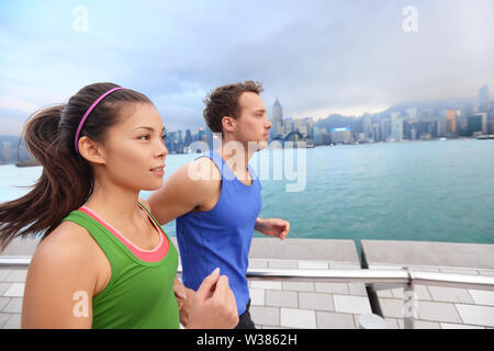 Running couple jogging in Hong Kong city. Runners training on Tsim Sha Tsui Promenade and Avenue of Stars in Victoria harbour, Kowloon, Hong Kong. Fitness runner man and sport woman model working out. Stock Photo