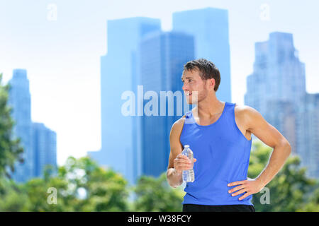 Sport man drinking water bottle in New York City. Male runner sweaty and thirsty after run in Central Park, NYC, Manhattan, with urban buildings skyline in the background.