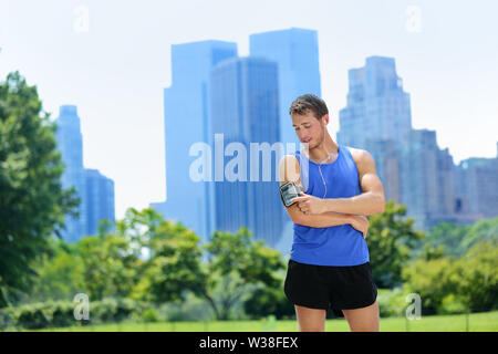 New York City man runner listening music on smartphone. Male adult jogger running using touchscreen on armband for workout in Central Park with urban background of Manhattan's skyscrapers skyline. Stock Photo