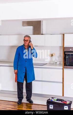 Mature handyman is standing in the kitchen near to the toolbox, talking on mobile and looking happy Stock Photo