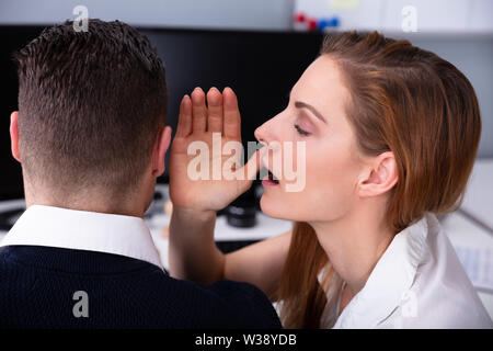 Close-up Of Young Businesswoman Whispering Secret Into Male Colleague's Ear In Office Stock Photo