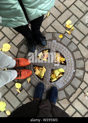 Luke with the inscription Sewerage of Leningrad. Top view of the pavement tile with autumn leaves. Feet in the shoes of two women and one man. St. Pet Stock Photo