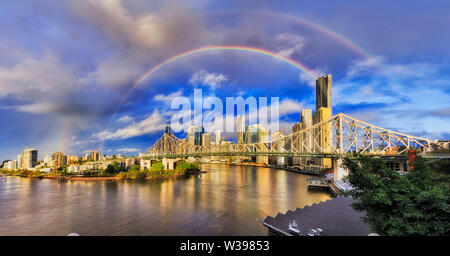 Story bridge across Brisbane river in front of Brisbane city CBD during morning afer rain with double rainbows over the city landmarks in blue sky. Stock Photo