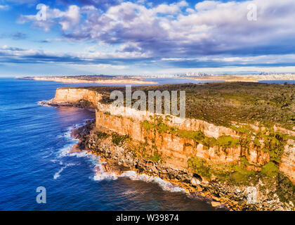 Steep sandstone cliffs of Sydney North Head at the entrance to Sydney harbour from Pacific ocean on a sunny morning with distant city CBD towers on th Stock Photo