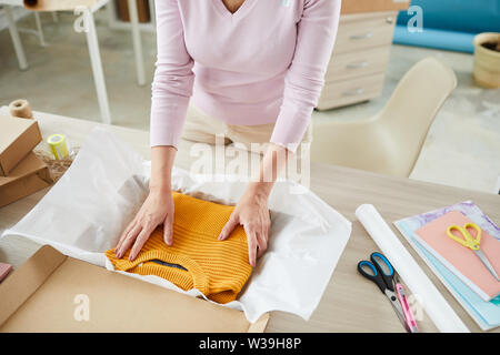 Young female employee putting folded yellow sweater on paper and in box while packing parcel Stock Photo