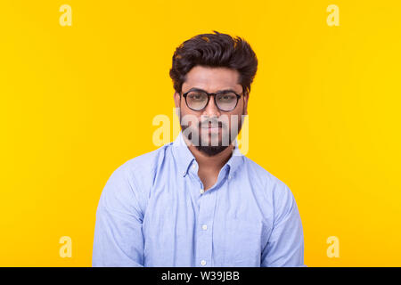 Young indian male professor with a beard in glasses and formal clothes is looking down thoughtfully posing on a yellow background. Concept of search Stock Photo