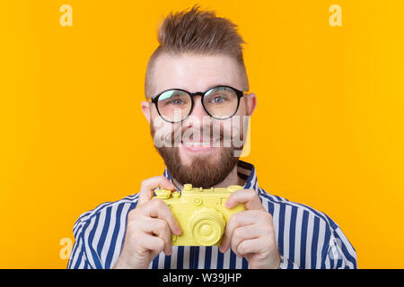 Cute young male photographer with a mustache and beard is photographing a yellow vintage camera on a yellow background. Concept of hobby and Stock Photo