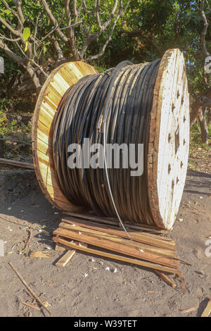Large wooden coil with electric cable laid in the nature on boards and black volcanic sand. The sun's rays illuminate the object. Tropical jungle Stock Photo