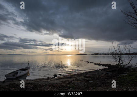 Beautiful view of Trasimeno lake shore (Umbria) at sunset, with a little boat, skeletal tree and sun coming down Stock Photo