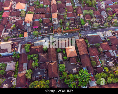 Many villas with brown-orange shingle roofs between tropical trees on the sky background in Ubud on Bali. Sun is shining onto them. Aerial horizontal