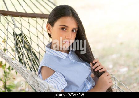 Attractive serious young girl with long dark hair wearing a stylish summer outfit relaxing outdoors in the garden in a hammock in the shade on a hot s Stock Photo