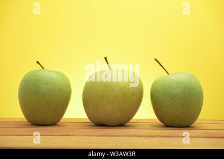 Three green apples on a red wooden table, yellow background. Stock Photo