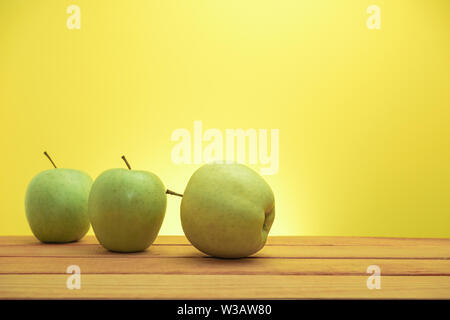 Three green apples on a red wooden table, yellow background. Stock Photo